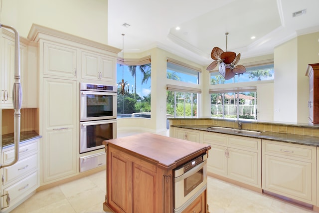 kitchen with a raised ceiling, hanging light fixtures, ceiling fan, dark stone countertops, and stainless steel double oven