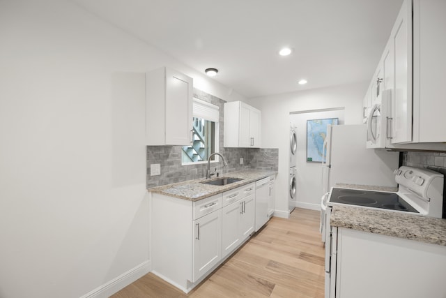 kitchen with light stone counters, white appliances, sink, stacked washer / dryer, and white cabinetry