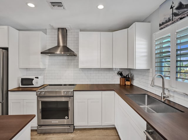 kitchen with appliances with stainless steel finishes, sink, butcher block counters, white cabinetry, and wall chimney exhaust hood