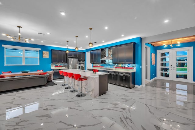 kitchen with french doors, a kitchen island with sink, wall chimney range hood, decorative light fixtures, and appliances with stainless steel finishes