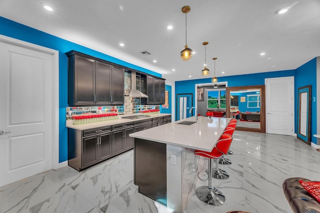 kitchen featuring decorative backsplash, hanging light fixtures, a kitchen island with sink, wall chimney exhaust hood, and sink