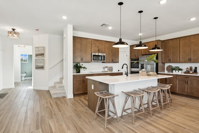 kitchen featuring an island with sink, hanging light fixtures, stainless steel appliances, a kitchen bar, and light wood-type flooring