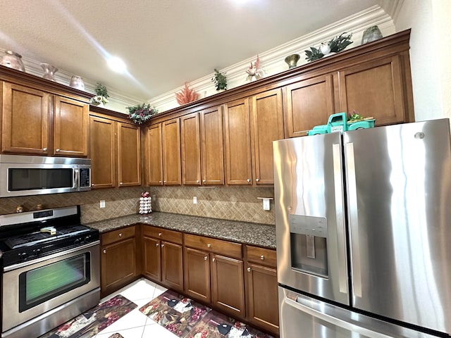 kitchen featuring stainless steel appliances, crown molding, dark stone counters, decorative backsplash, and light tile patterned flooring