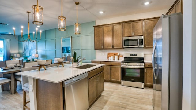 kitchen with an island with sink, stainless steel appliances, sink, decorative light fixtures, and light wood-type flooring