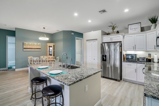 kitchen with stainless steel fridge, white cabinets, a kitchen island with sink, light wood-type flooring, and decorative light fixtures