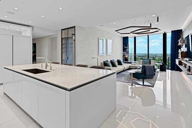 kitchen featuring white cabinetry, an island with sink, sink, a wall of windows, and light stone countertops