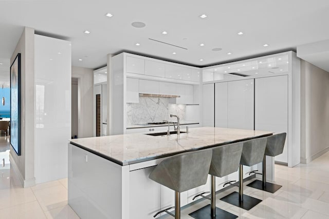 kitchen with a kitchen island with sink, light stone counters, tasteful backsplash, white cabinets, and a kitchen bar