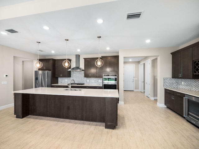 kitchen featuring appliances with stainless steel finishes, light wood-type flooring, wall chimney range hood, a center island with sink, and hanging light fixtures