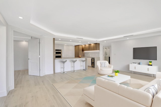 living room with wine cooler, sink, a tray ceiling, and light wood-type flooring