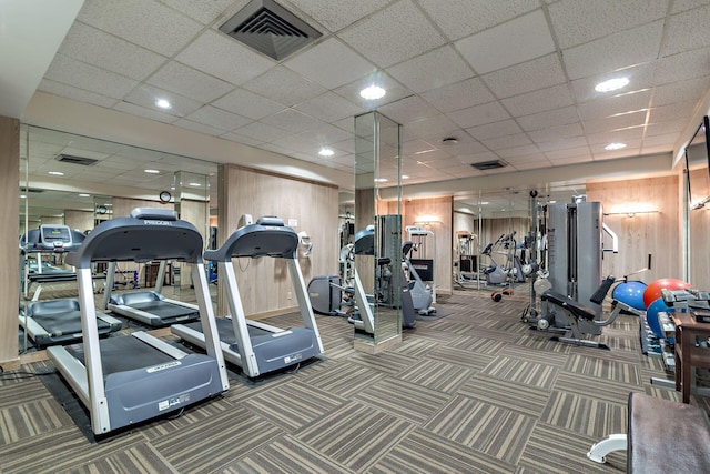 gym with a paneled ceiling, wooden walls, and dark carpet