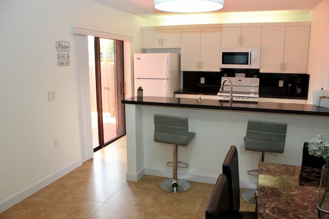 kitchen featuring backsplash, dark stone countertops, a breakfast bar, white cabinetry, and white appliances