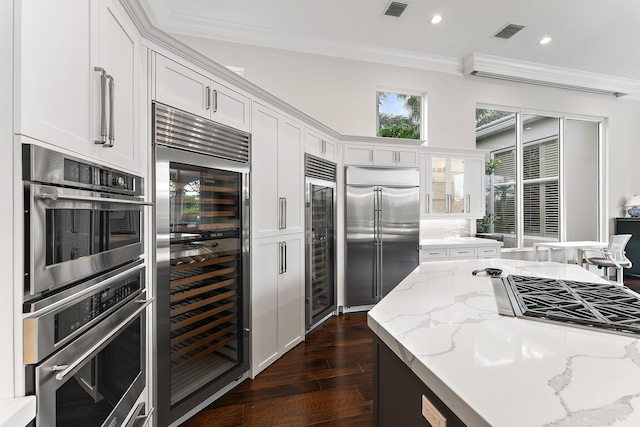 kitchen with light stone countertops, ornamental molding, stainless steel appliances, beverage cooler, and white cabinetry