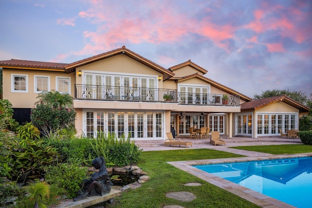 back house at dusk featuring french doors, a balcony, ceiling fan, a patio, and a lawn