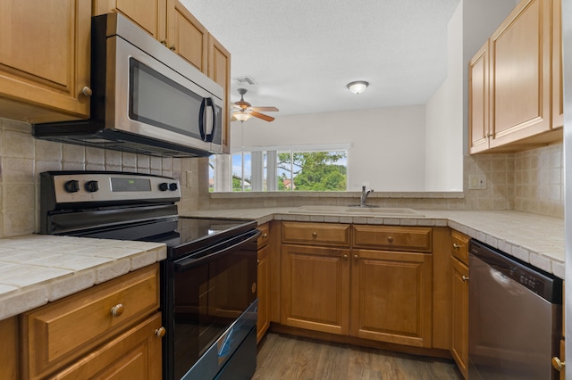 kitchen with tasteful backsplash, ceiling fan, appliances with stainless steel finishes, tile countertops, and light hardwood / wood-style floors