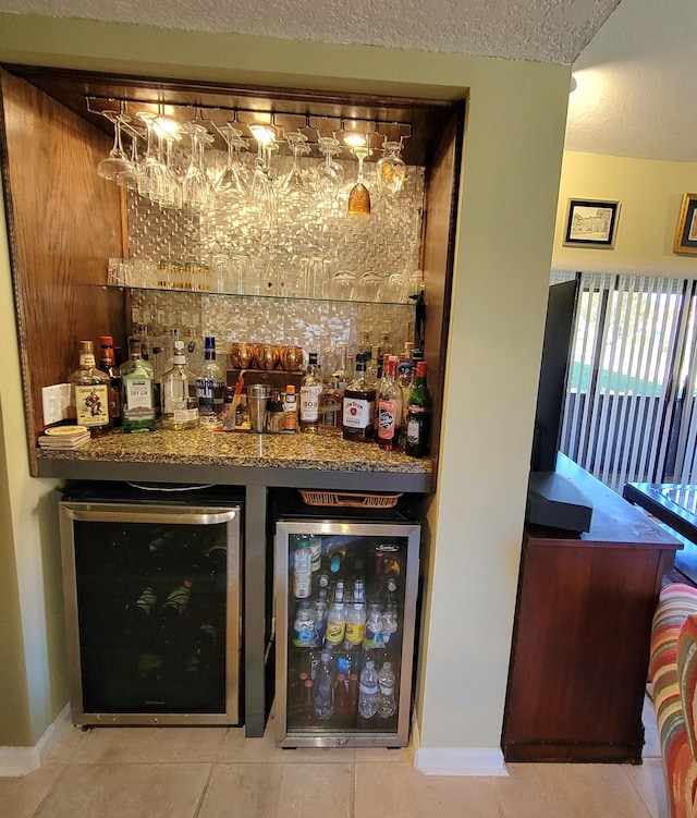 bar with a textured ceiling, light tile patterned floors, dark stone counters, and beverage cooler