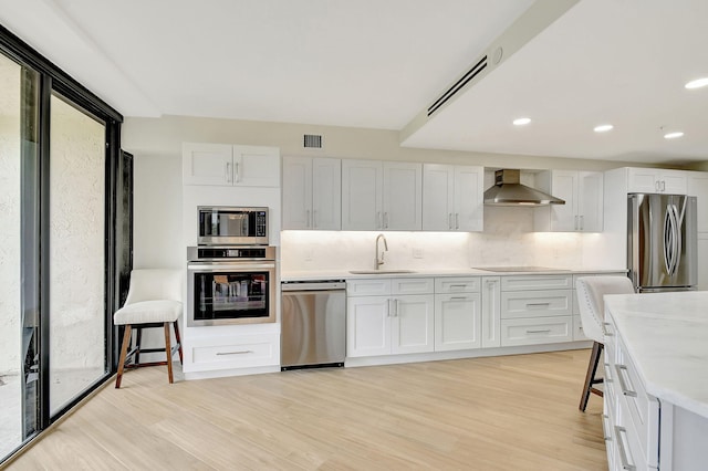 kitchen featuring wall chimney range hood, sink, white cabinetry, appliances with stainless steel finishes, and light hardwood / wood-style floors