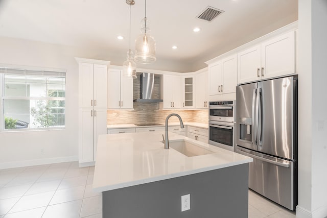 kitchen with stainless steel appliances, wall chimney exhaust hood, hanging light fixtures, sink, and an island with sink