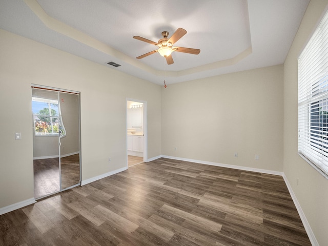 unfurnished bedroom with ensuite bathroom, ceiling fan, dark hardwood / wood-style floors, a tray ceiling, and a closet