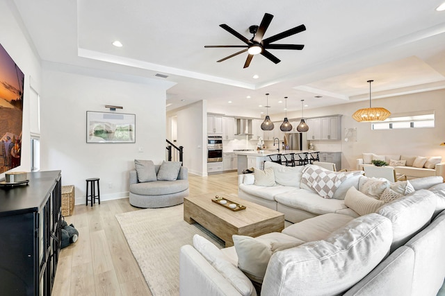 living room featuring a raised ceiling, ceiling fan, sink, and light wood-type flooring