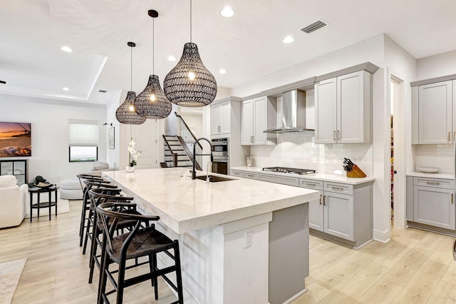 kitchen with light stone countertops, wall chimney exhaust hood, light hardwood / wood-style floors, a center island with sink, and gray cabinets