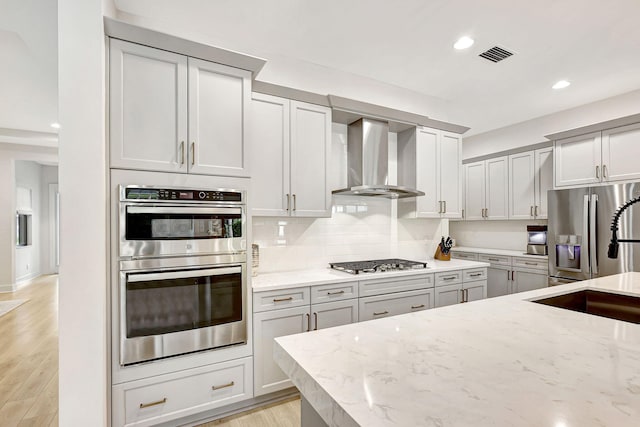 kitchen with light stone countertops, backsplash, wall chimney exhaust hood, stainless steel appliances, and light hardwood / wood-style floors