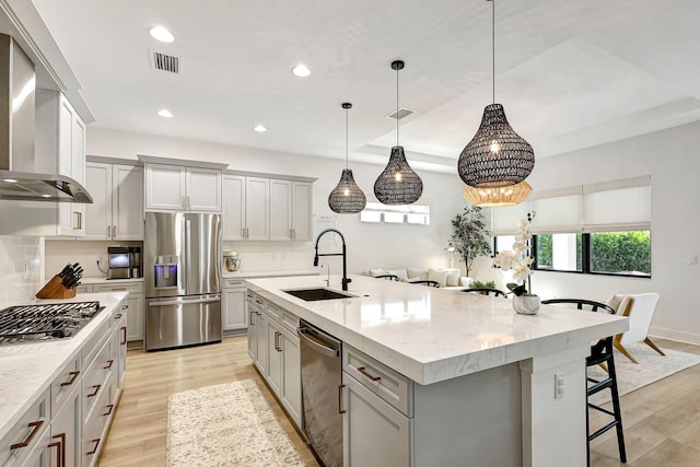 kitchen featuring a center island with sink, wall chimney range hood, sink, appliances with stainless steel finishes, and decorative light fixtures