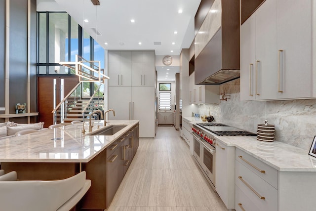 kitchen with double oven range, wall chimney range hood, white cabinetry, pendant lighting, and a kitchen island with sink