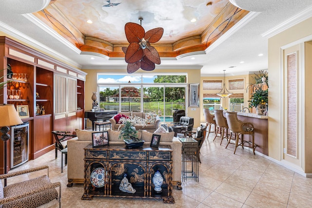 tiled living area featuring a tray ceiling, crown molding, ceiling fan, a dry bar, and baseboards
