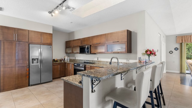 kitchen with a textured ceiling, kitchen peninsula, a kitchen breakfast bar, stainless steel appliances, and dark stone counters