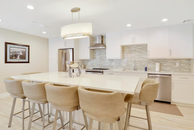 kitchen featuring pendant lighting, white cabinets, wall chimney range hood, light wood-type flooring, and stainless steel appliances