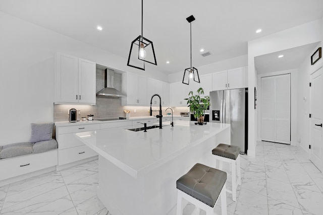 kitchen featuring an island with sink, stainless steel fridge with ice dispenser, white cabinets, wall chimney range hood, and pendant lighting