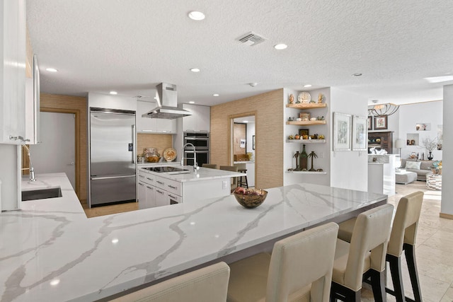 kitchen with a textured ceiling, white cabinetry, stainless steel appliances, wall chimney exhaust hood, and a center island with sink