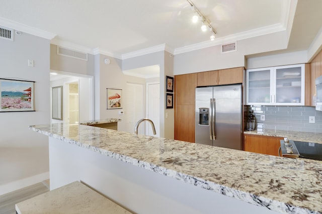 kitchen with stainless steel appliances, brown cabinetry, light stone counters, crown molding, and glass insert cabinets