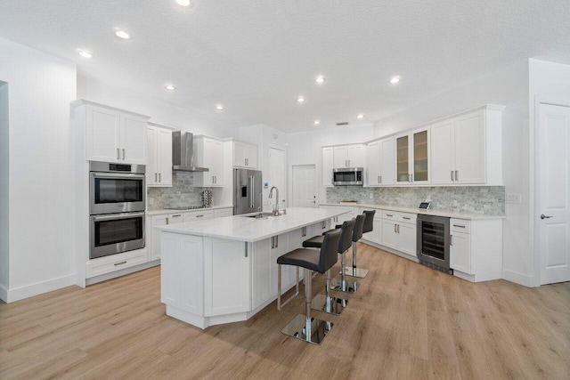 kitchen featuring stainless steel appliances, wine cooler, an island with sink, light hardwood / wood-style flooring, and wall chimney range hood