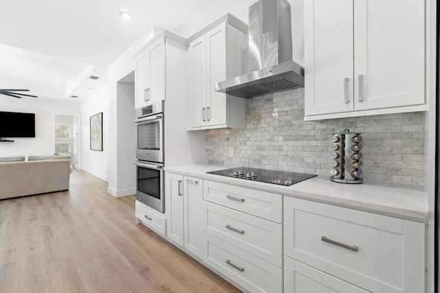 kitchen with black electric cooktop, wall chimney exhaust hood, stainless steel double oven, white cabinetry, and light wood-type flooring