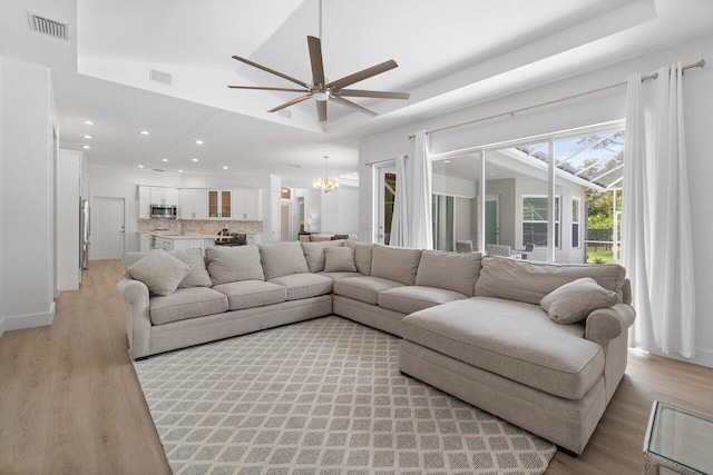 living room featuring light wood-type flooring, a raised ceiling, and ceiling fan with notable chandelier