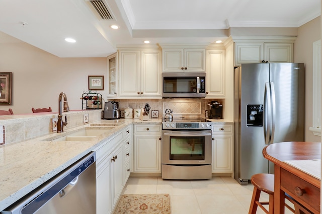 kitchen with stainless steel appliances, sink, cream cabinets, light stone countertops, and crown molding