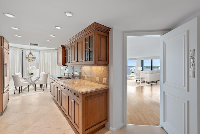 kitchen with light stone countertops, a sink, decorative backsplash, brown cabinets, and glass insert cabinets