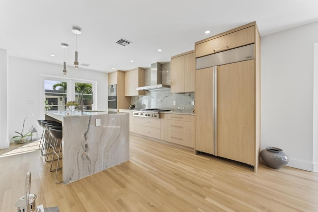 kitchen featuring wall chimney range hood, light brown cabinets, paneled built in fridge, light hardwood / wood-style floors, and an island with sink