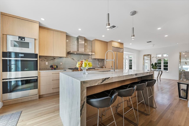 kitchen with a center island with sink, hanging light fixtures, light hardwood / wood-style flooring, wall chimney exhaust hood, and appliances with stainless steel finishes