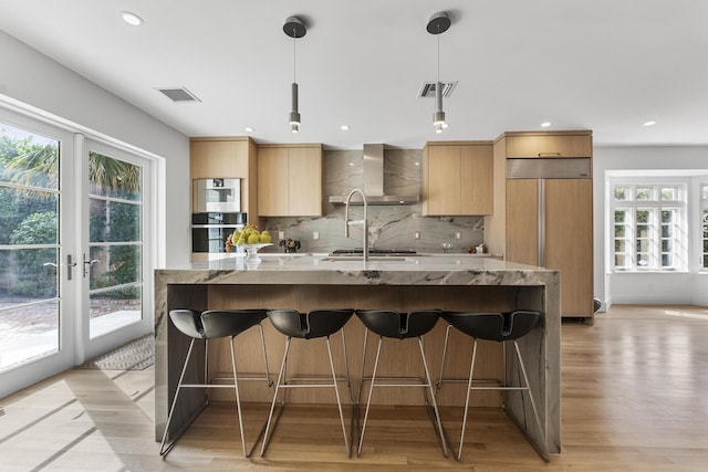 kitchen with pendant lighting, wall chimney range hood, light hardwood / wood-style flooring, light stone countertops, and stainless steel appliances