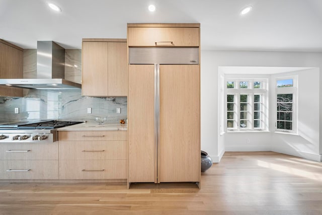 kitchen featuring wall chimney exhaust hood, paneled fridge, stainless steel gas stovetop, light brown cabinetry, and light wood-type flooring