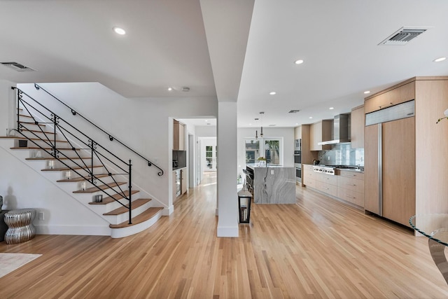 kitchen featuring backsplash, double oven, wall chimney range hood, light brown cabinets, and light hardwood / wood-style floors