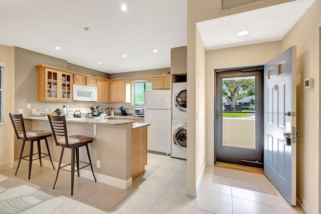 kitchen featuring kitchen peninsula, light brown cabinets, a kitchen bar, white appliances, and stacked washer / dryer