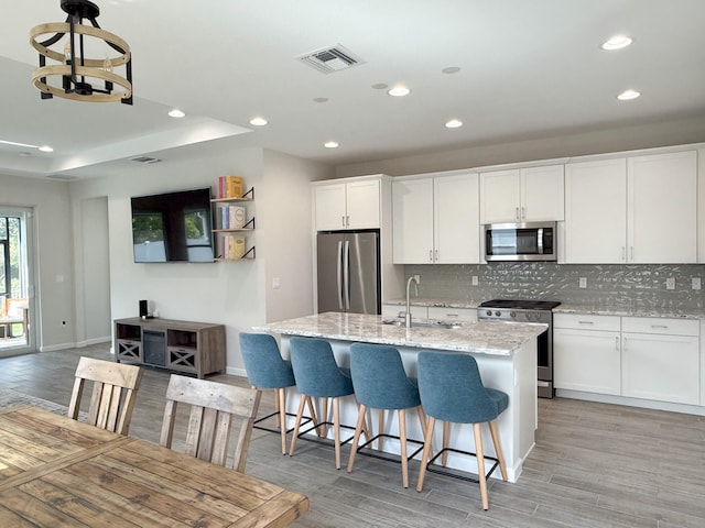 kitchen featuring light stone counters, stainless steel appliances, light hardwood / wood-style floors, white cabinetry, and an island with sink