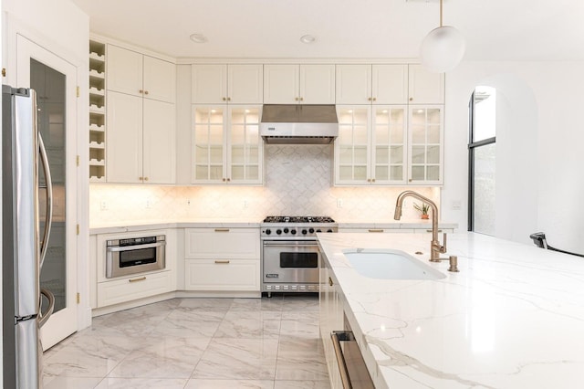 kitchen featuring stainless steel appliances, range hood, sink, light stone countertops, and decorative light fixtures