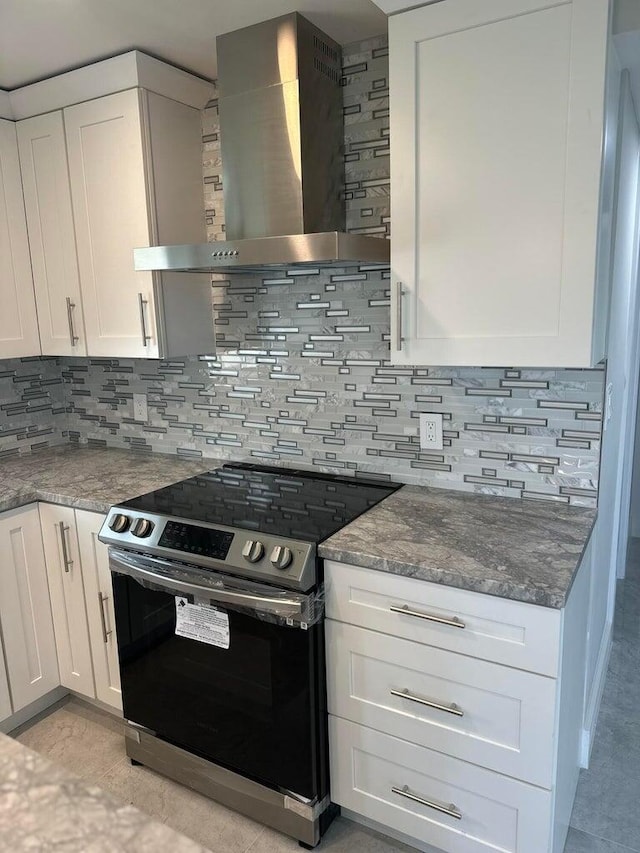 kitchen featuring stainless steel electric range, dark stone counters, white cabinets, wall chimney exhaust hood, and decorative backsplash