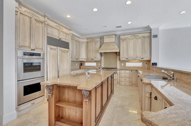 kitchen with sink, custom range hood, light stone countertops, stainless steel double oven, and a kitchen island with sink