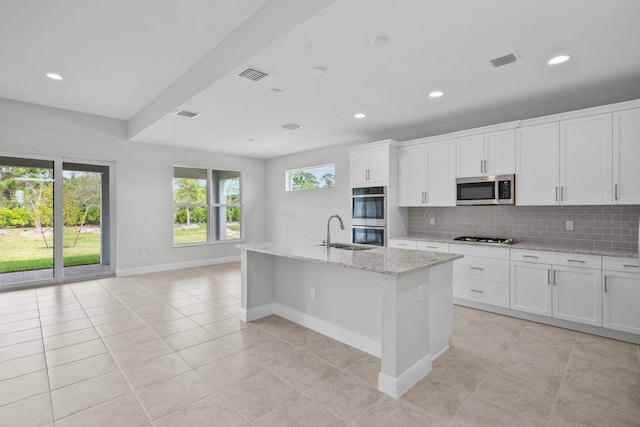 kitchen with appliances with stainless steel finishes, white cabinets, light stone counters, decorative backsplash, and a center island with sink