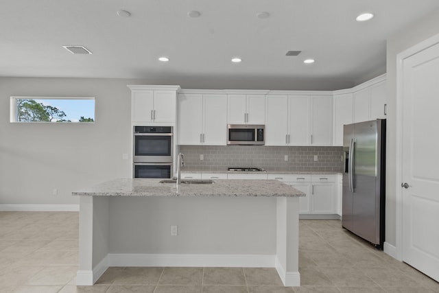 kitchen featuring tasteful backsplash, light stone countertops, appliances with stainless steel finishes, white cabinetry, and a center island with sink